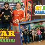 Three people standing side by side at Animasia Studio — from left to right, an Animasia representative in a pink shirt, a special Hollywood guest holding an Animasia 20th Anniversary tote bag, and another Animasia team member in an orange jersey. They are smiling in front of a vibrant wall filled with Malaysian animated characters, with the text “Hollywood’s Star Shines at Animasia!” and “#1 Happy Family USA.”